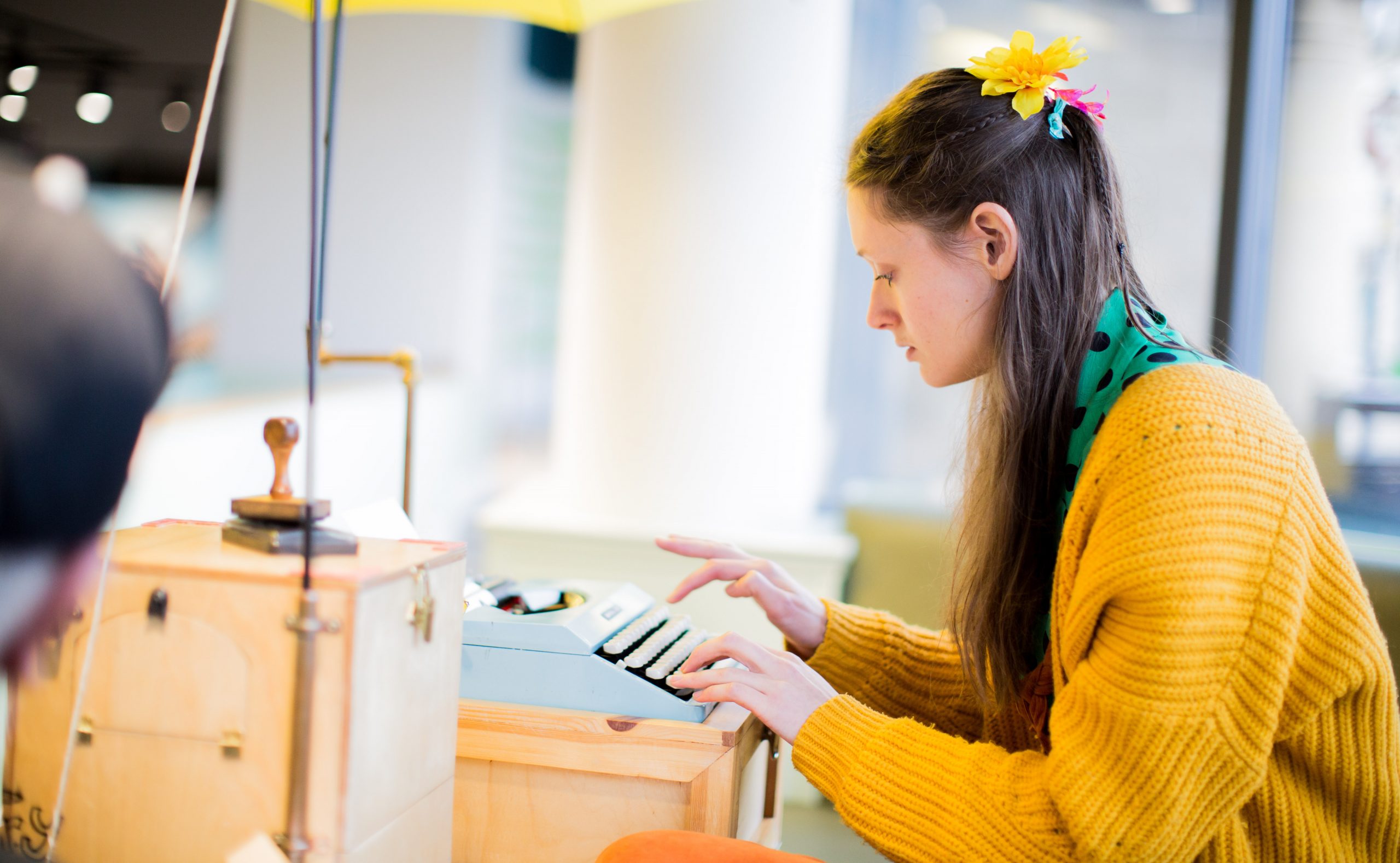 A woman in a yellow jumper with flowers in her hair types on a typewriter