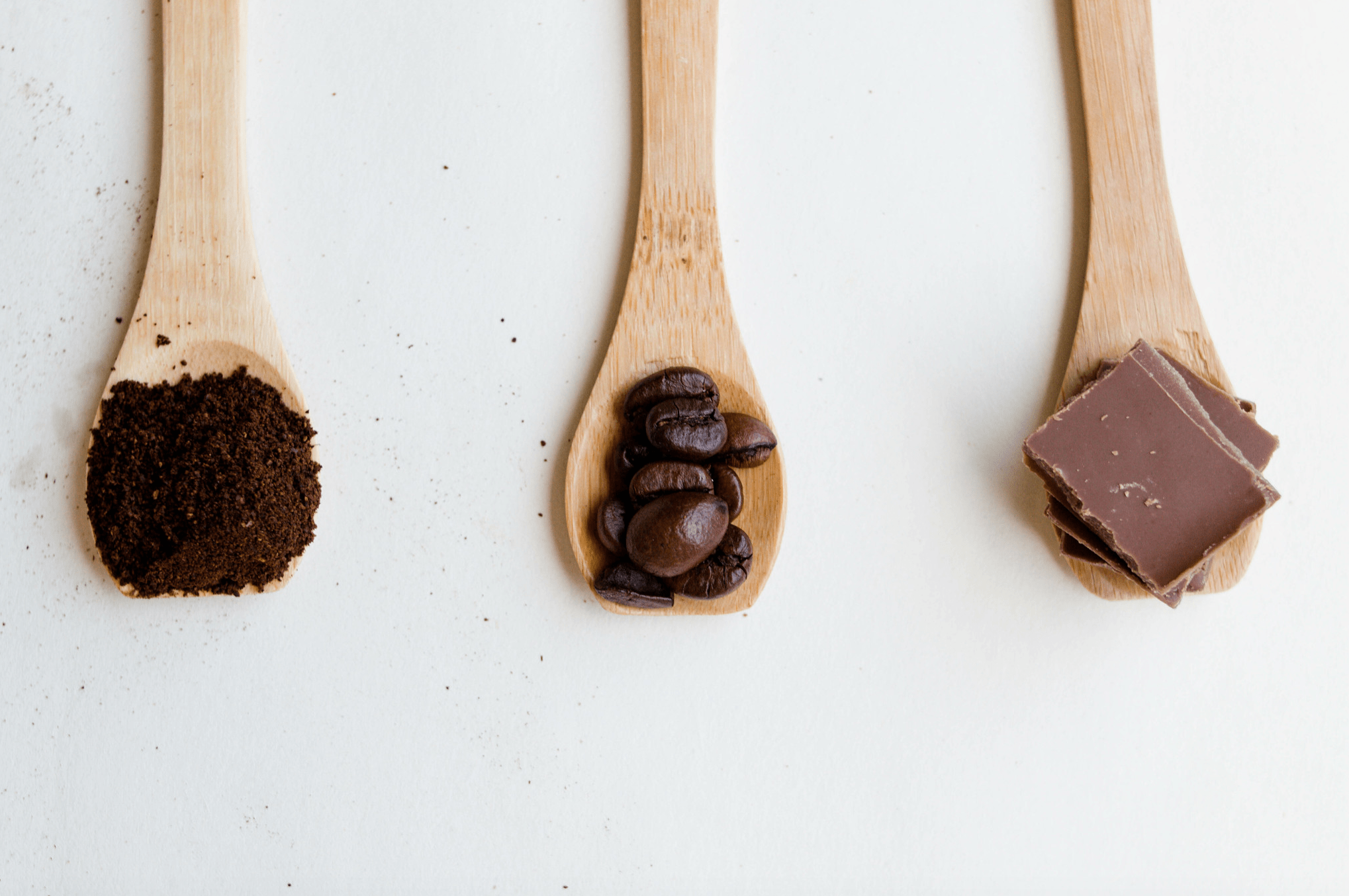 Three wooden spoons, one holding coffee granules, one coffee beans and one chocolate.
