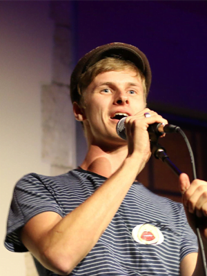 A man in striped t shirt and brown hat talking into a microphone on stage