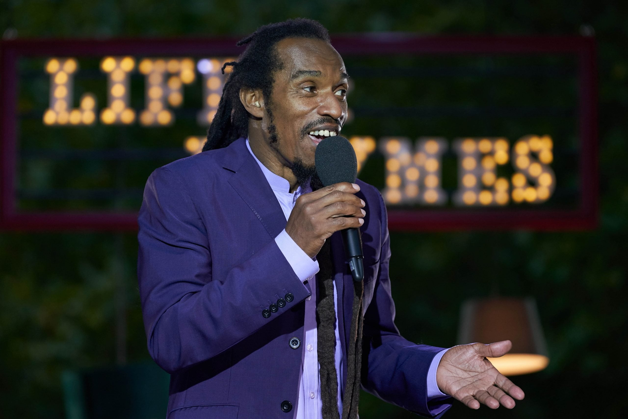 Photo of a man speaking into a microphone with dreadlocks in a ponytail and a blue suit