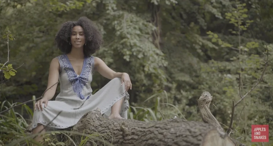 Screenshot of 'With You I See Light', a poetry film by Aisha Sanyang-Meek. It shows Aiysha with her hair loose, sitting in a forest. Her pose is strong.