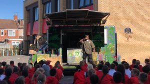 A man dressed in green holds a mic to his mouth standing in front of a decorated van, performing to school children