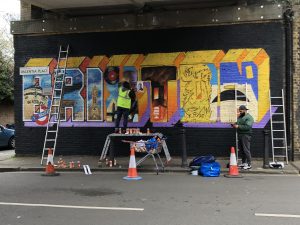 Two artists paint a mural of Brixton on a wall in an underpass.