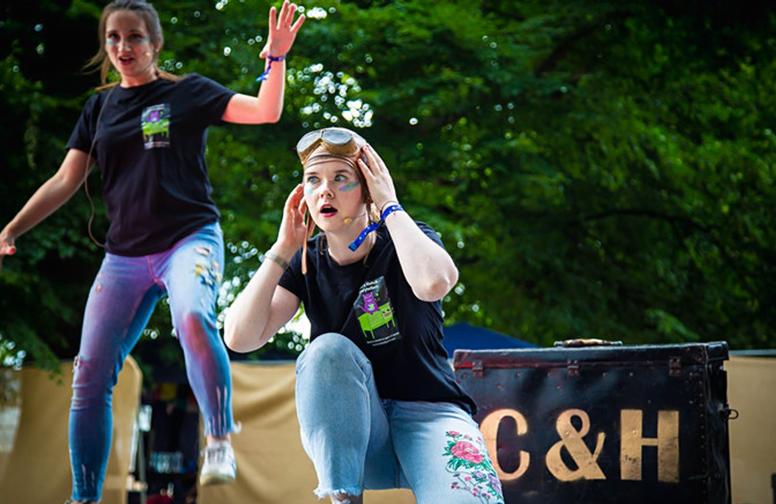 Two women on an outdoor stage, acting shocked with hands in the air