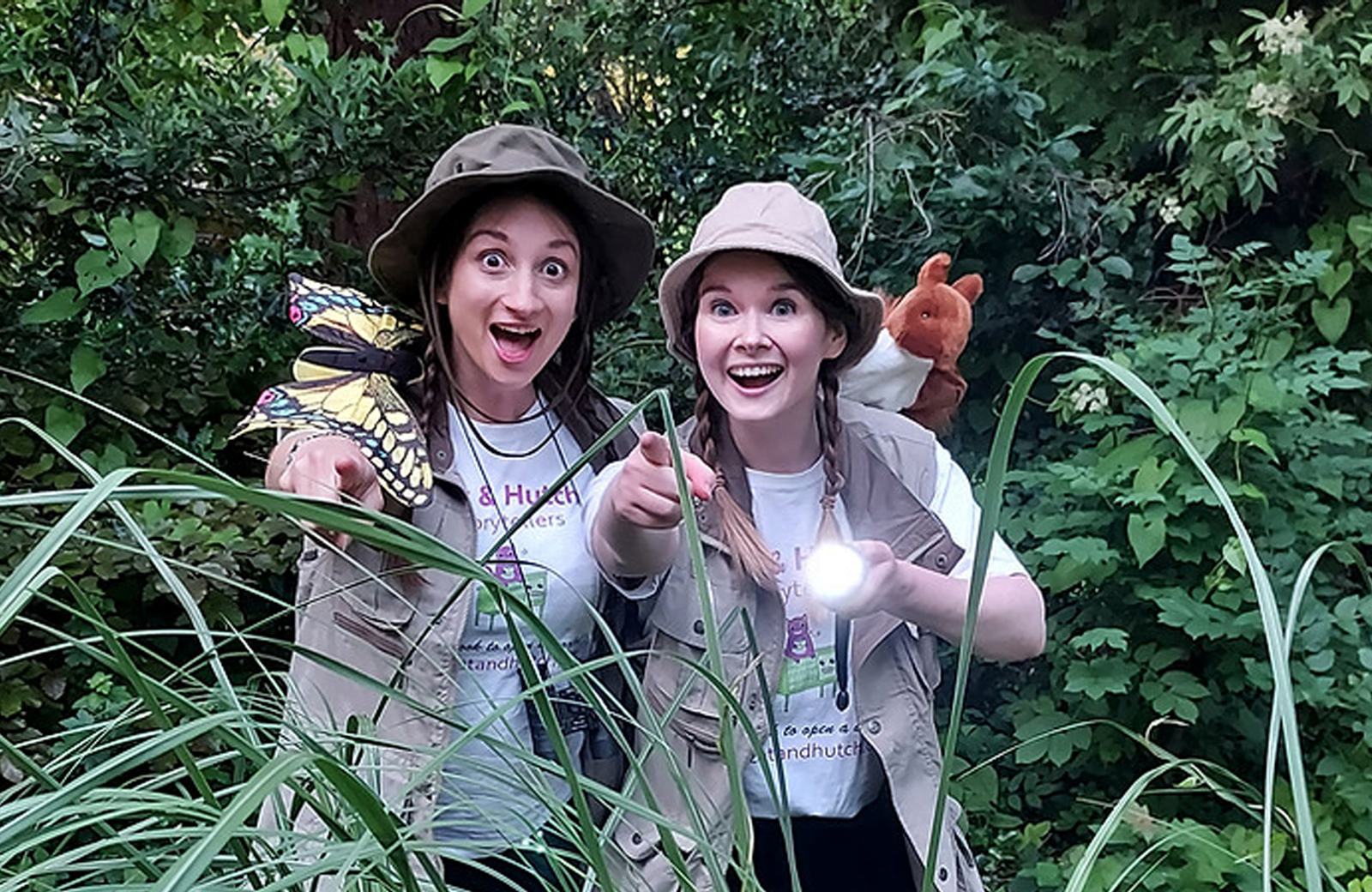 Two women in safari outfits amongst leaves looking bewildered, one with a large butterfly on her arm
