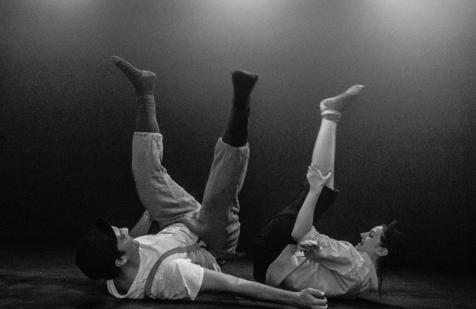 Two people on their bands with knees in the air, smiling, wearing bowler hats. Photo is in black and white, like a comedy classic