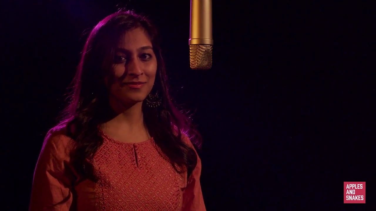 Screenshot from a video of Bhumika standing next to a microphone in a dark room with pink lighting