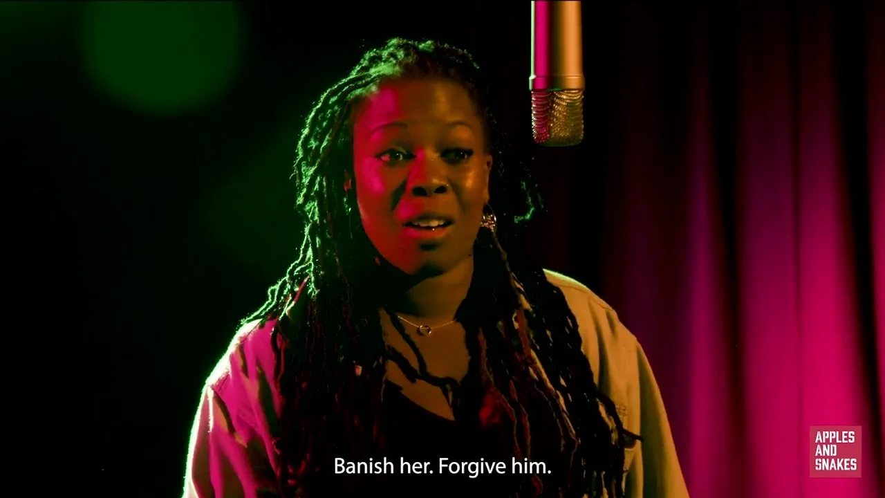 Screenshot from a video of Desree speaking into a microphone in a dark room with green yellow and pink lighting