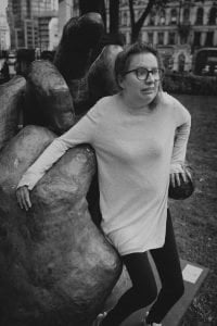 Black and white image of Helen wearing glasses, a pale coloured tunic and leggings, leaning against a statue of a hand