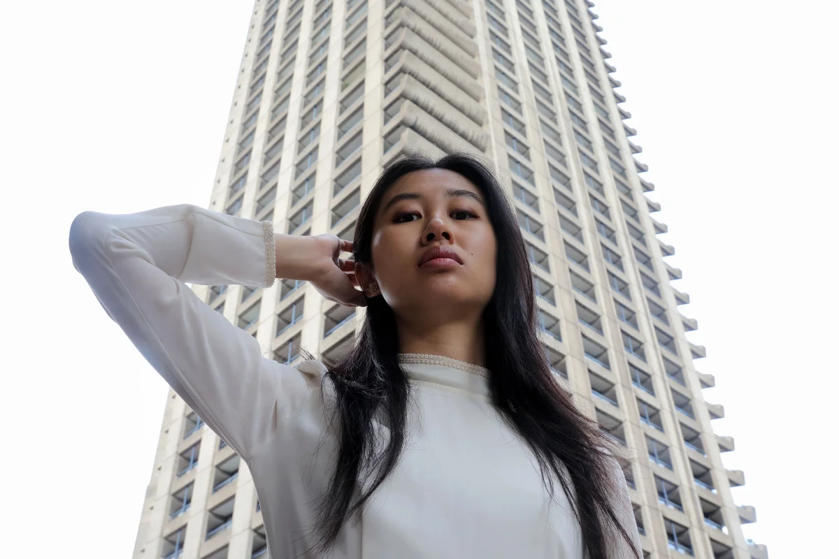 A woman with long dark hair wearing a high necked, long sleeve white dress puts her arm behind her head, standing in front of a tall tower block