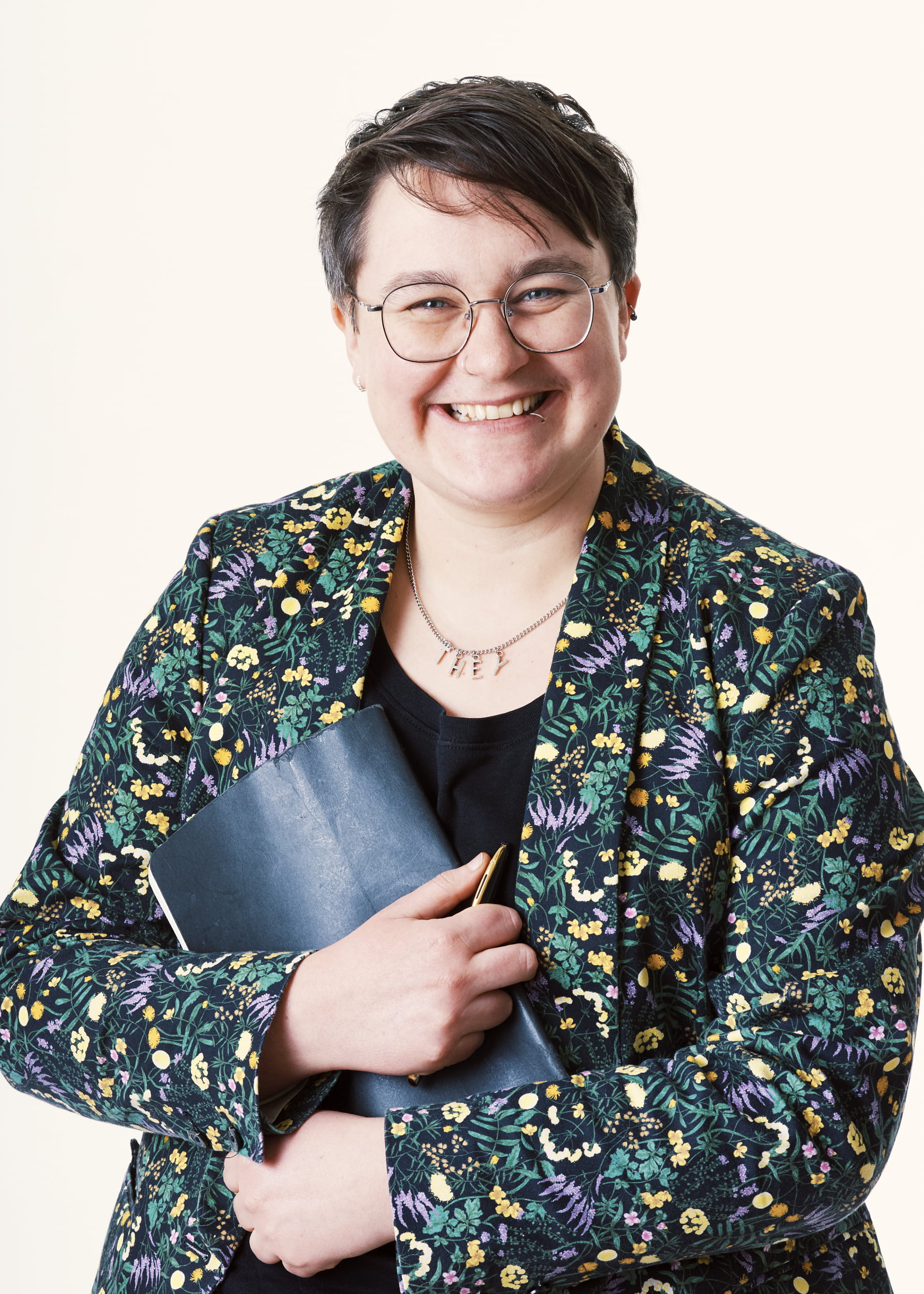 Lindon smiles, holding a leather bound book. Short hair, round glasses and wearing a patterned colourful blazer and black t-shirt
