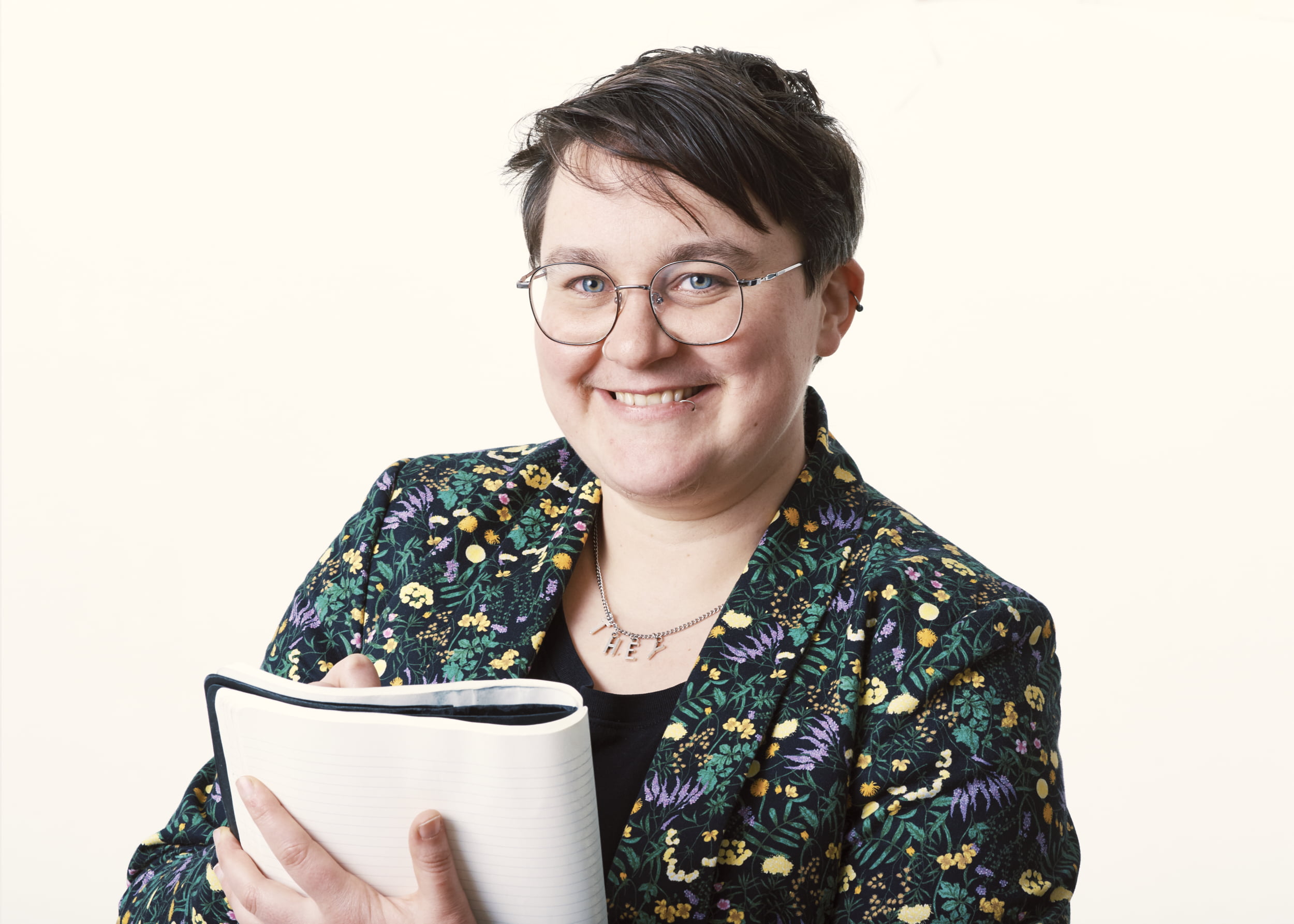 Lindon smiles, holding a leather bound book. Short hair, round glasses and wearing a patterned colourful blazer and black t-shirt