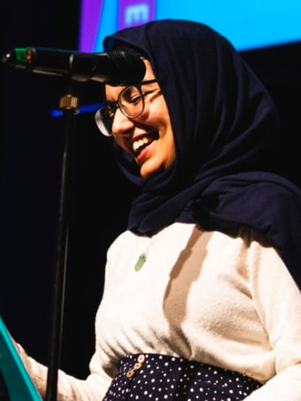 Aliyah wears dark head scarf, white cardigan top and polka dot skirt, smiling into a mic on stage