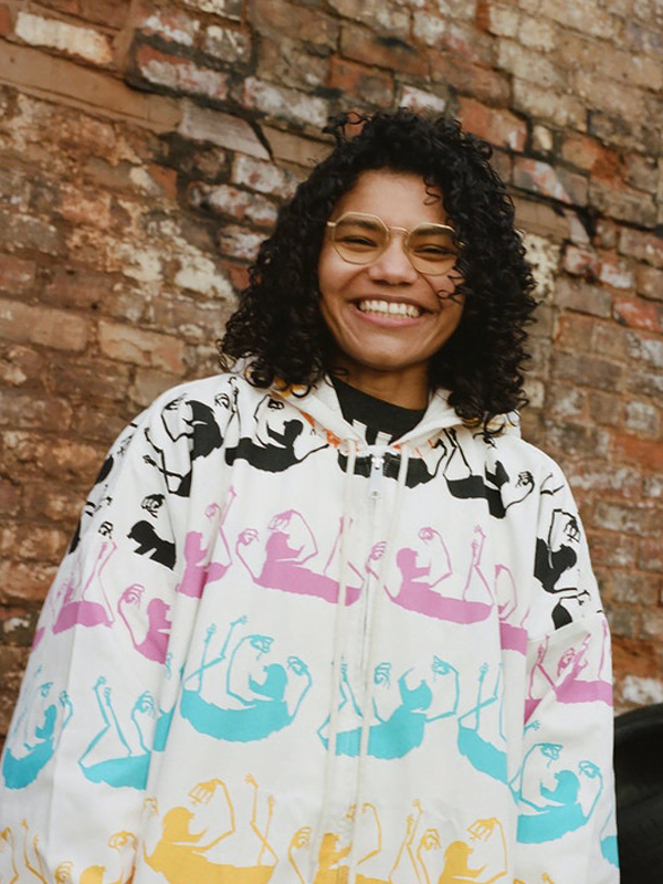 Ella wears a white hoodie with with black, yellow, pink and light blue sloths. Dark curly bob, with gold rimmed glasses