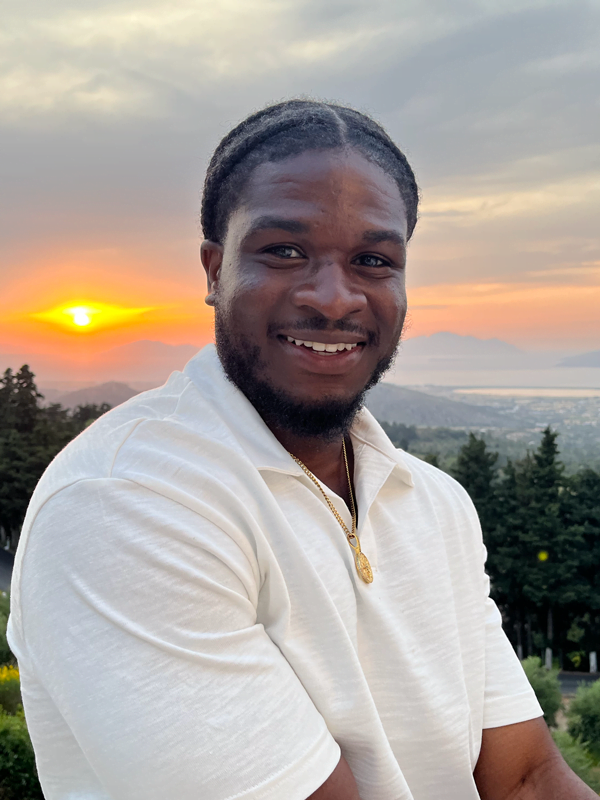 Garfar wears his hair braided and pulled back, and white polo type shirt with gold pendant necklace. Sunset in background against trees and mountain.