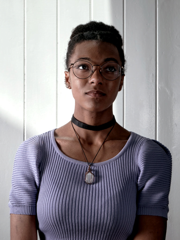 Ingrid has a piped violet top, black choker and gemstone necklace, against a white panel background