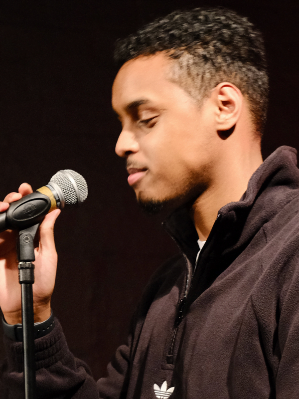 Zakariye (young adult) wearing a black hoody with short black hair, about to talk into a mic