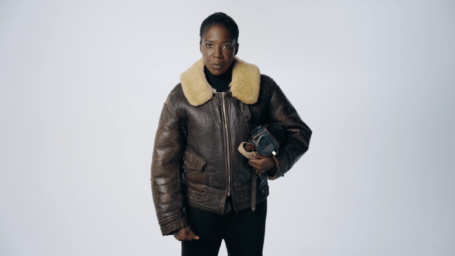Kat Francois stands holding a pilots helmet wearing a leather and fur aviator jacket