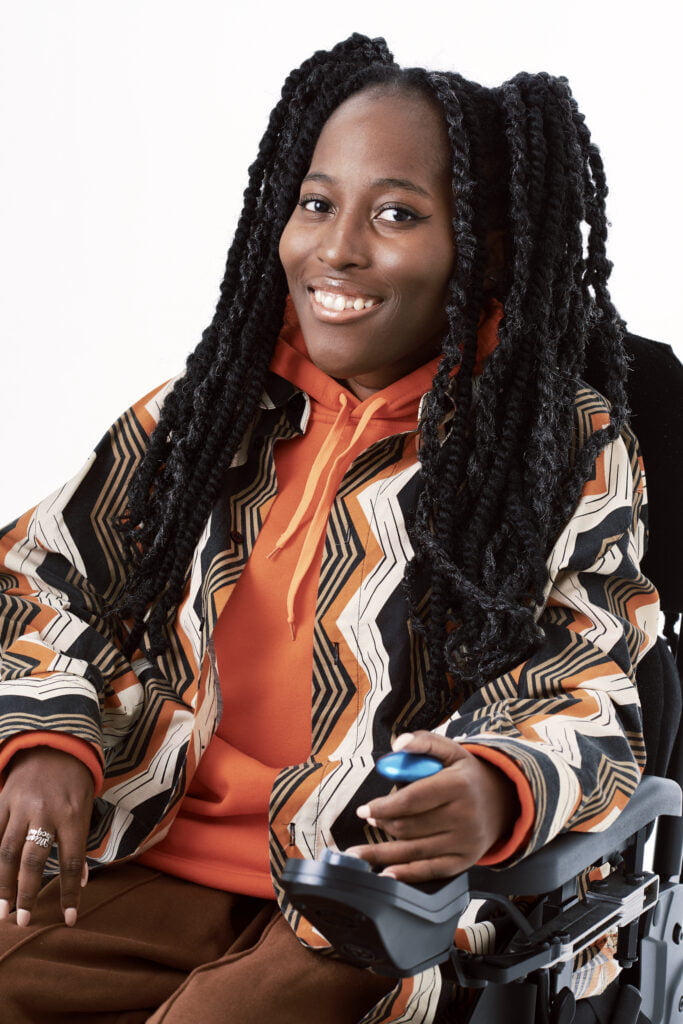 An image of poet Miss Jacqui. She has long black hair and smiles towards the camera. She wears an orange hoodie and an orange, white and black jacket