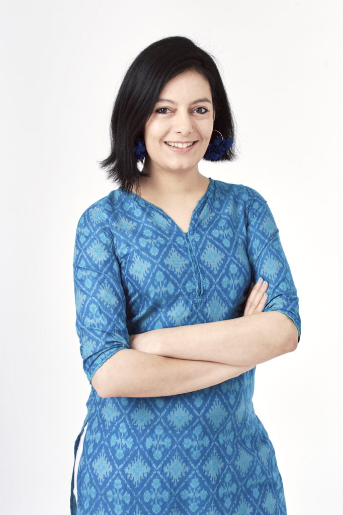 An image of poet Laila Sumpton. She has black shoulder length hair, she wears a blue patterned dress and crosse her arms. She smiles out towards the camera.