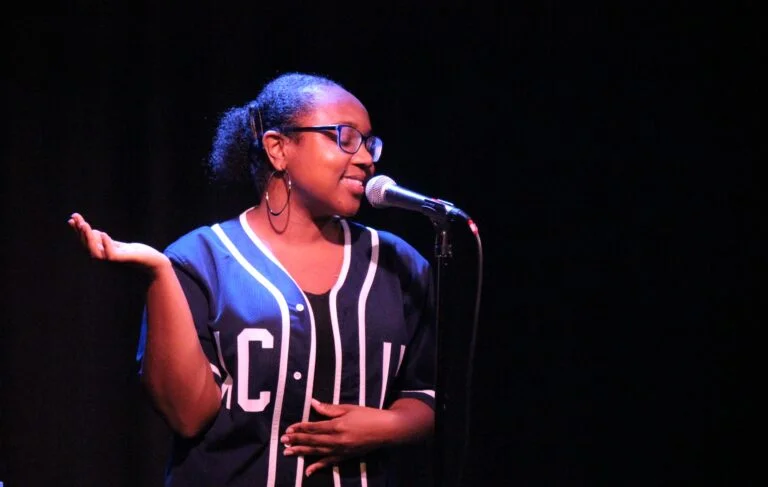 Poet Shaniqua Benjamin stands at a microphone. She wears black glasses, has hooped earrings and has shoulder length black hair tied back. Shaniqua wears a navy cardigan with white piping. She smiles as she performs.