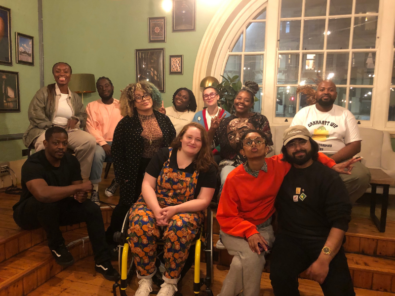 Mohoganny Brown sits smiling with a group of mixed adult poets in a dimly lit room